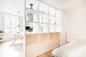A modern bedroom with a large bed, a built-in wardrobe with multiple shelves, a desk with a chair, and a large window providing natural light. - Chiado Studio Portuguese Charm with AC (Lisboa)