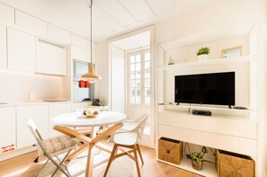 Eine moderne Küche mit weißen Schränken, einem runden Esstisch mit Stühlen, einer hängenden Pendelleuchte und einem an der Wand montierten Flachbildfernseher.