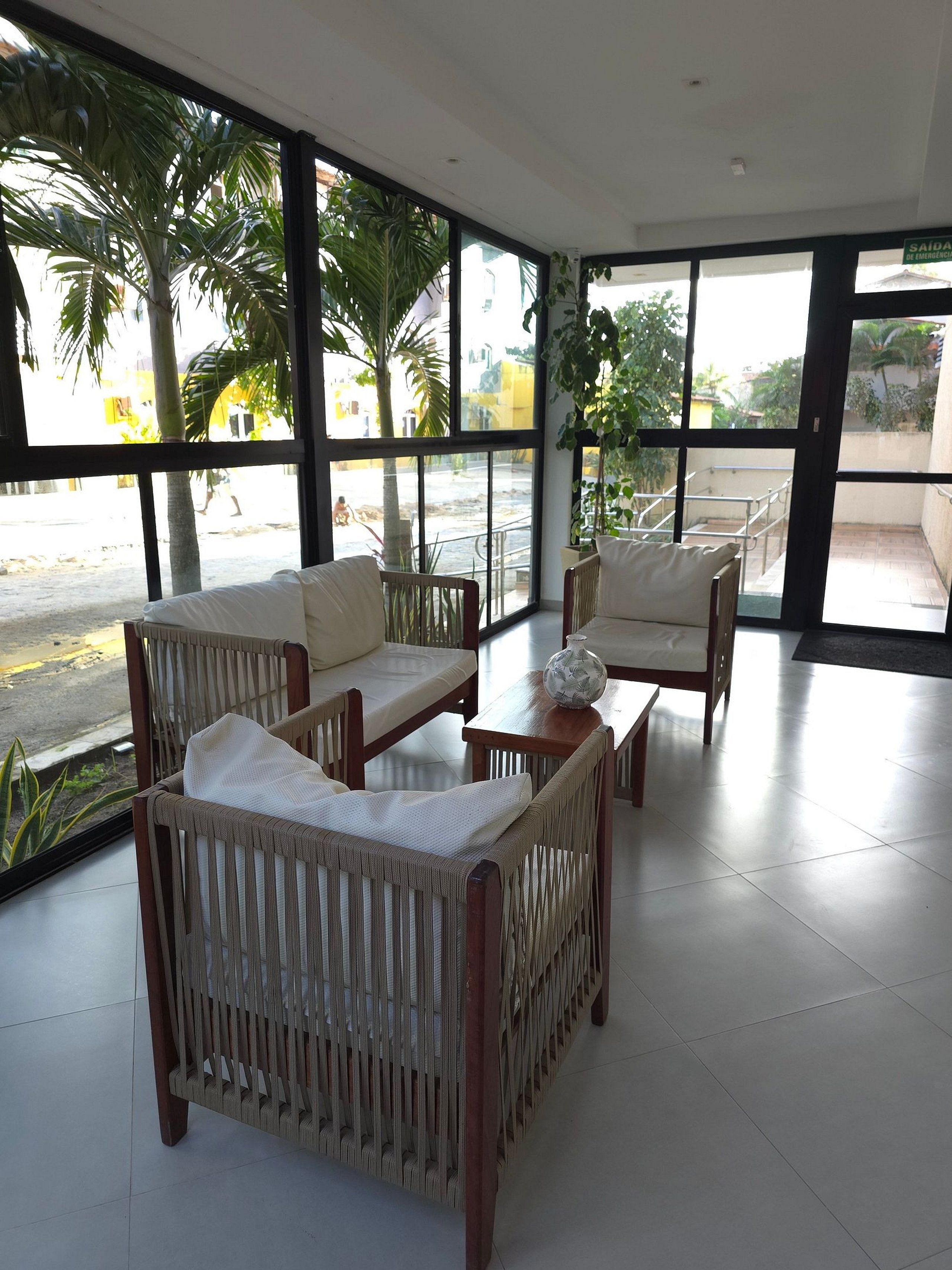 Amplia habitación con ventanas grandes, muebles de mimbre y vista a la calle y palmeras.