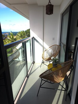 A balcony with two wicker chairs, a hanging light fixture, and a view of the ocean and buildings. - MB I - Ap. 405 - Nico Uruguay (Ipojuca)