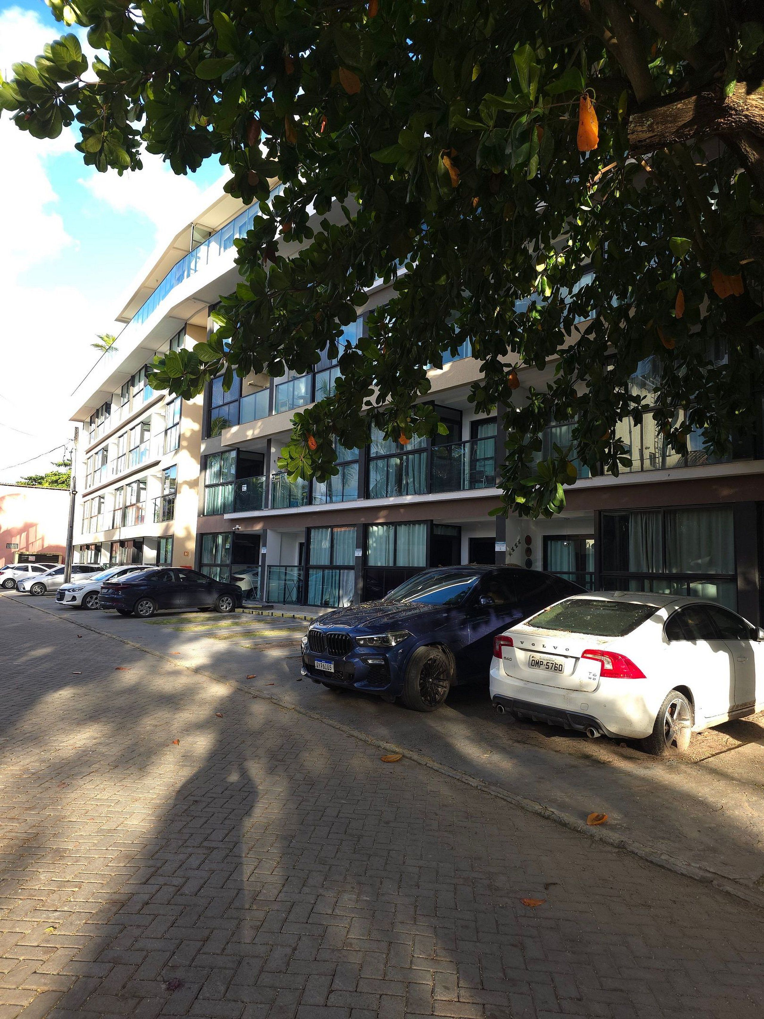 Un área de estacionamiento con varios automóviles, incluyendo un Volvo blanco y un SUV azul, frente a un edificio moderno con varios pisos y grandes ventanales.