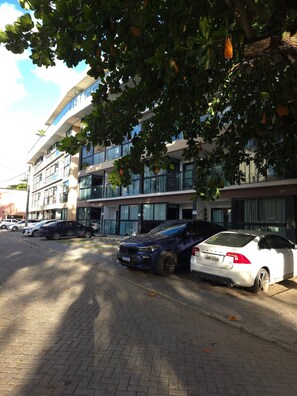 A parking area with several cars, including a white Volvo and a blue SUV, in front of a modern building with multiple floors and large windows. - MB I - Ap. 405 - Nico Uruguay (Ipojuca)