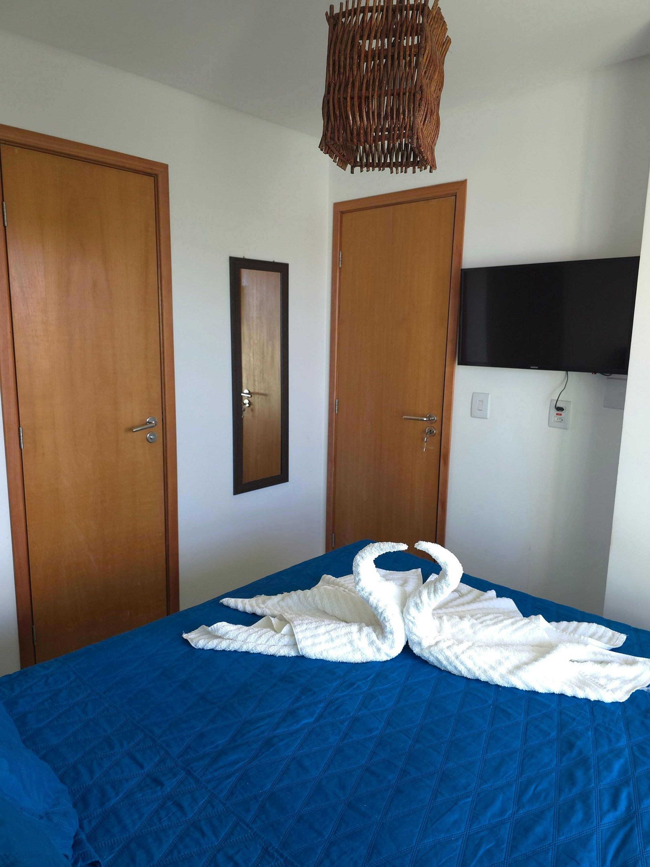 A bedroom with two wooden doors, a framed mirror, a flat-screen TV mounted on the wall, and a bed with a blue quilt and white towels arranged in a swan shape.