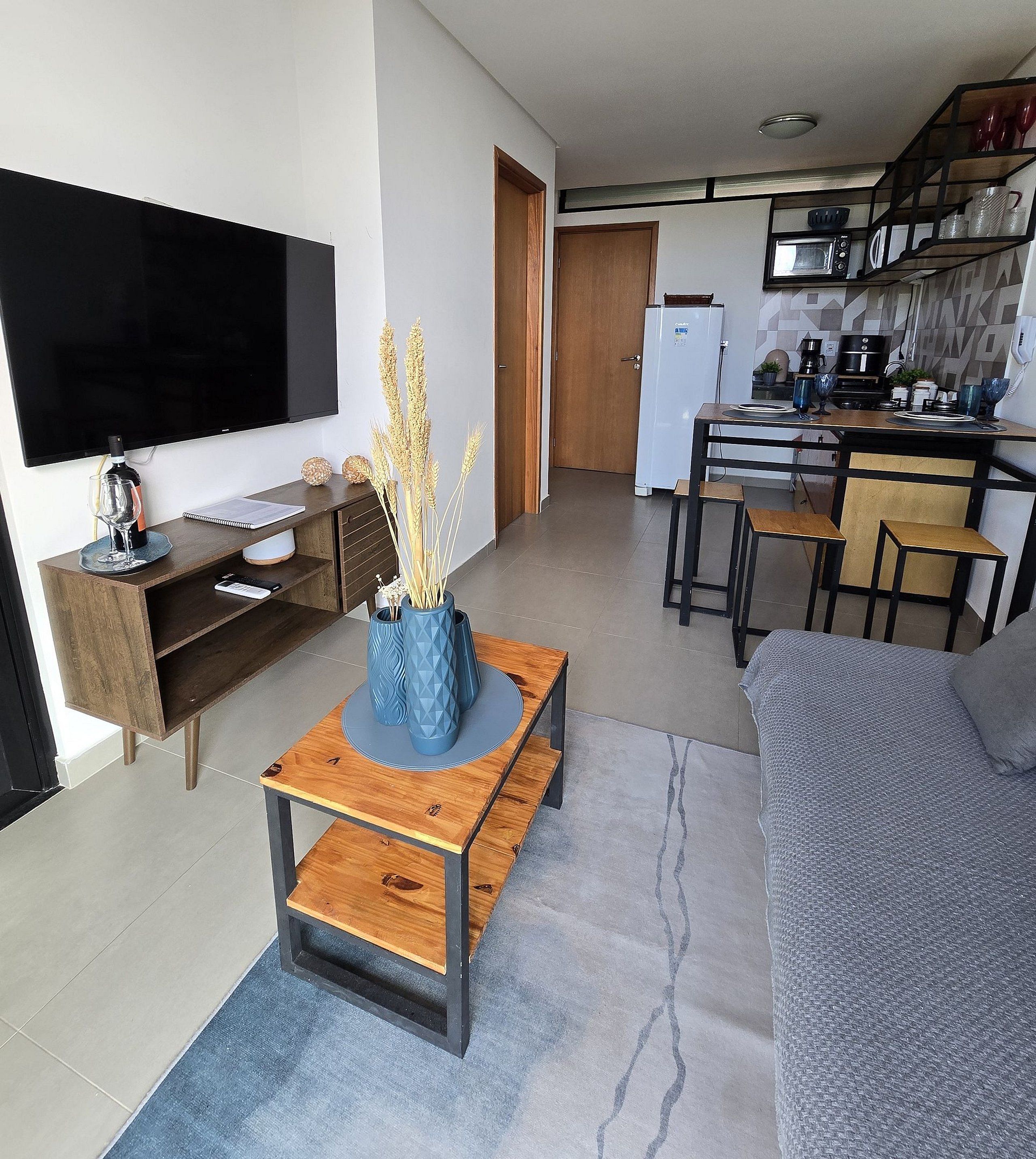Un salón moderno con un televisor de pantalla plana, una mesa de centro de madera con jarrones azules, un sofá gris y una zona de cocina con microondas y armarios.