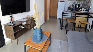 Un salón moderno con un televisor de pantalla plana, una mesa de centro de madera con jarrones azules, un sofá gris y una zona de cocina con microondas y armarios.