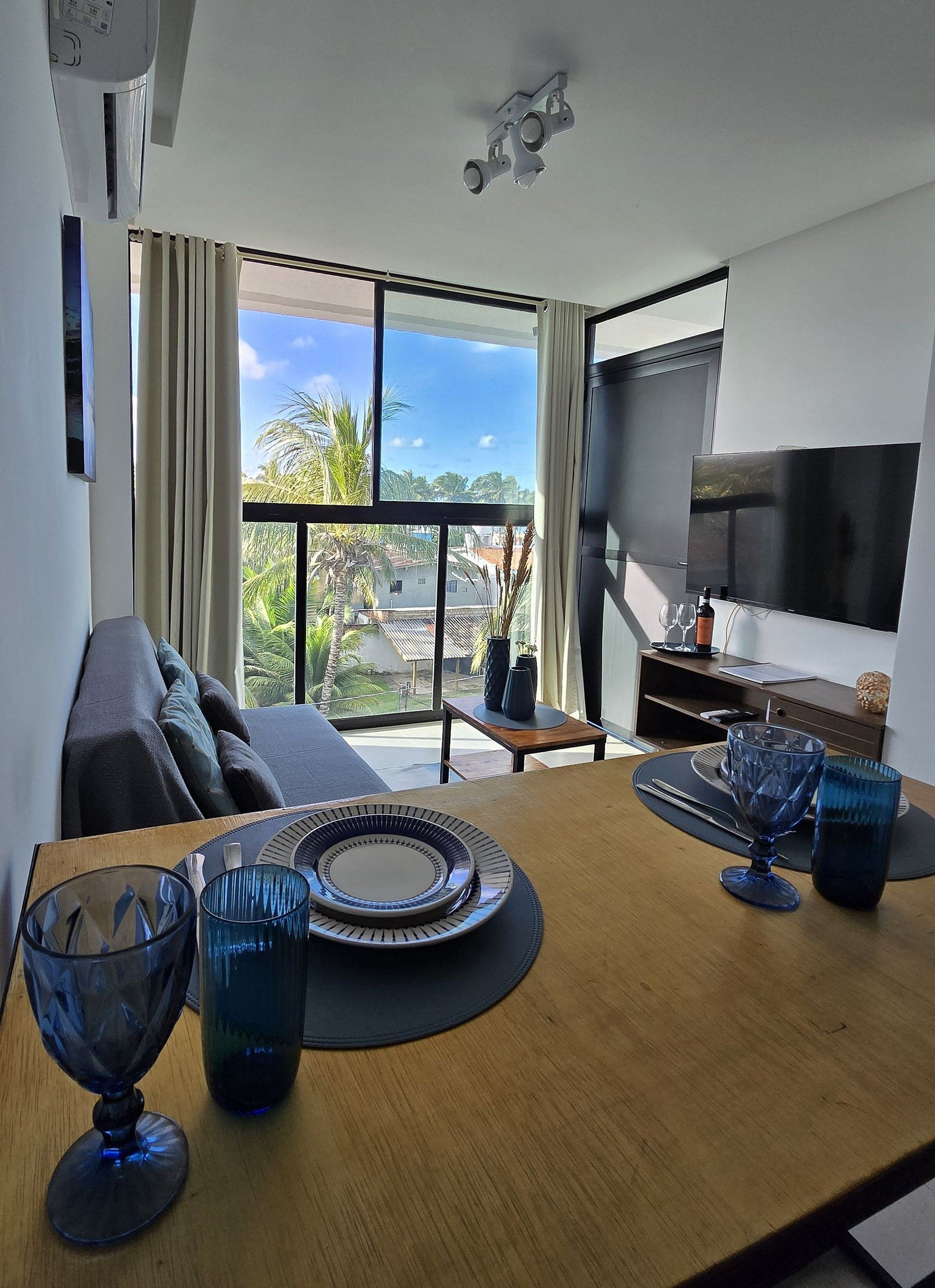 A modern living room with a large TV, a dining table with blue glassware, a sofa, and a view of palm trees and buildings.