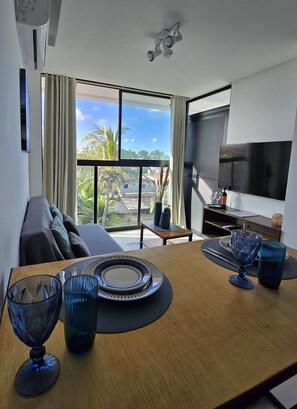 A modern living room with a large TV, a dining table with blue glassware, a sofa, and a view of palm trees and buildings. - MB I - Ap. 405 - Nico Uruguay (Ipojuca)