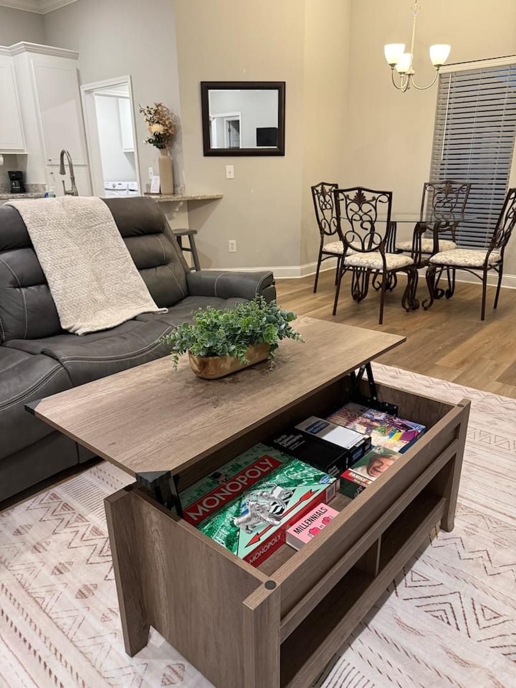 A living room with a sofa, a coffee table with a plant, a bookshelf with board games, and a dining area with chairs.