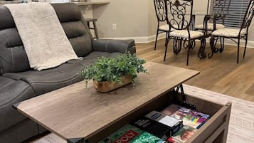 A living room with a sofa, a coffee table with a plant, a bookshelf with board games, and a dining area with chairs.