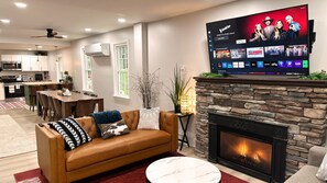 A modern living room with a leather sofa, a flat-screen TV mounted above a stone fireplace, and a dining area with a wooden table and chairs. - 3-BR Chalet in Lake Harmony - Lakeview, 6 Queen Beds, Fireplace, Walk to Town (Lake Harmony)
