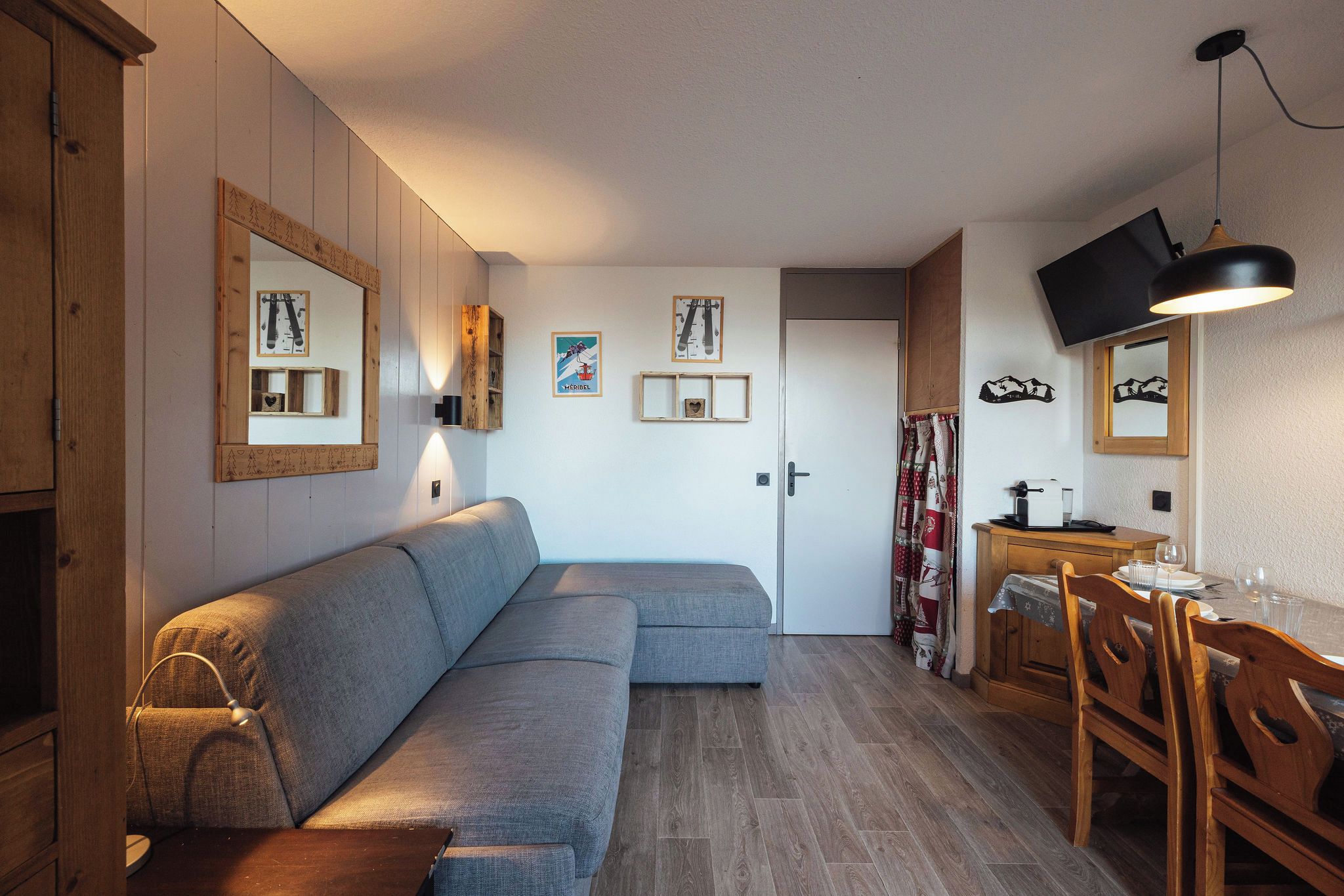 A cozy living area with a sofa, wooden cabinets, a dining table with chairs, and a television mounted on the wall.