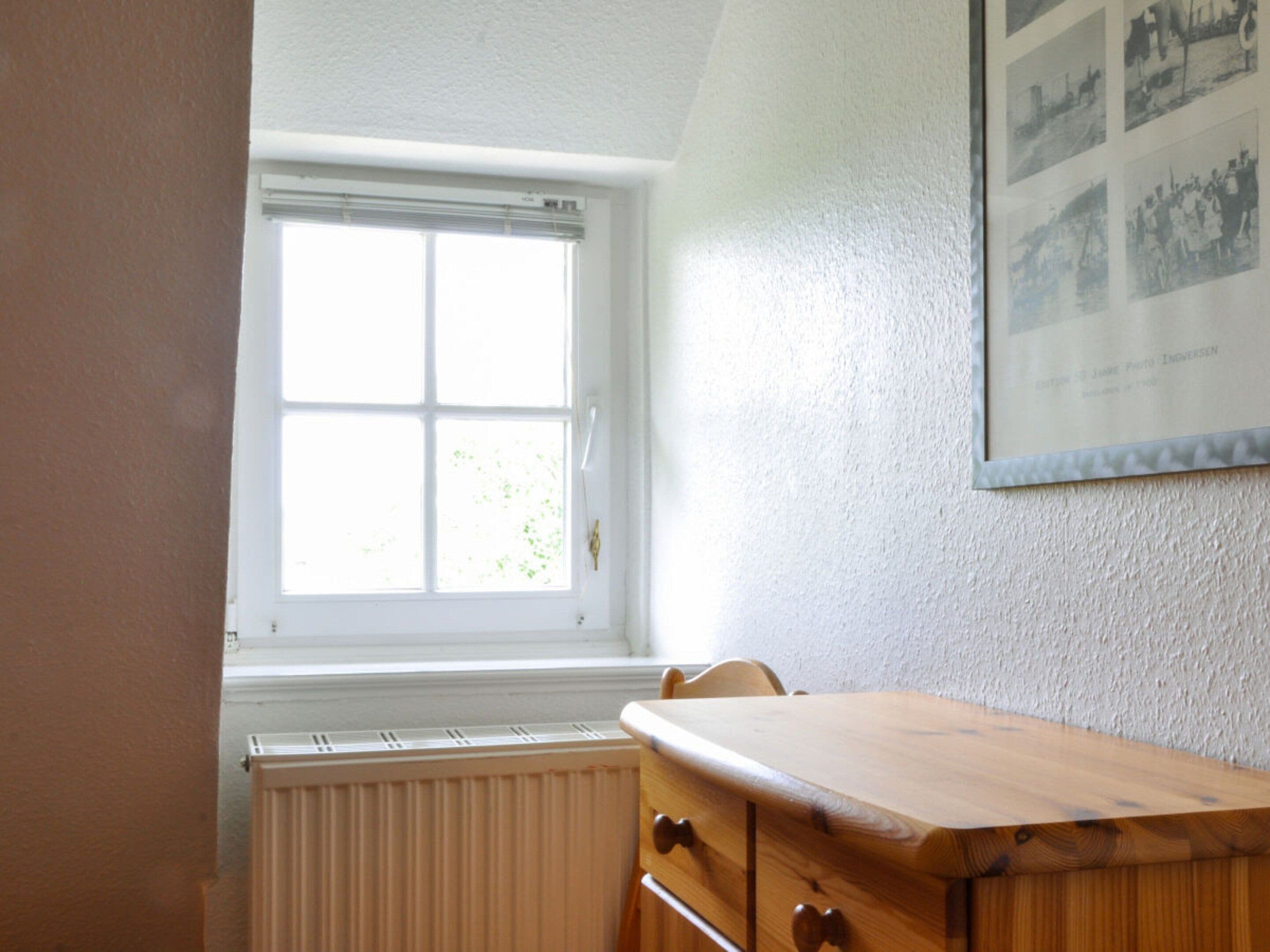 A room with a window, a radiator, a wooden table, and a wall-mounted photo.