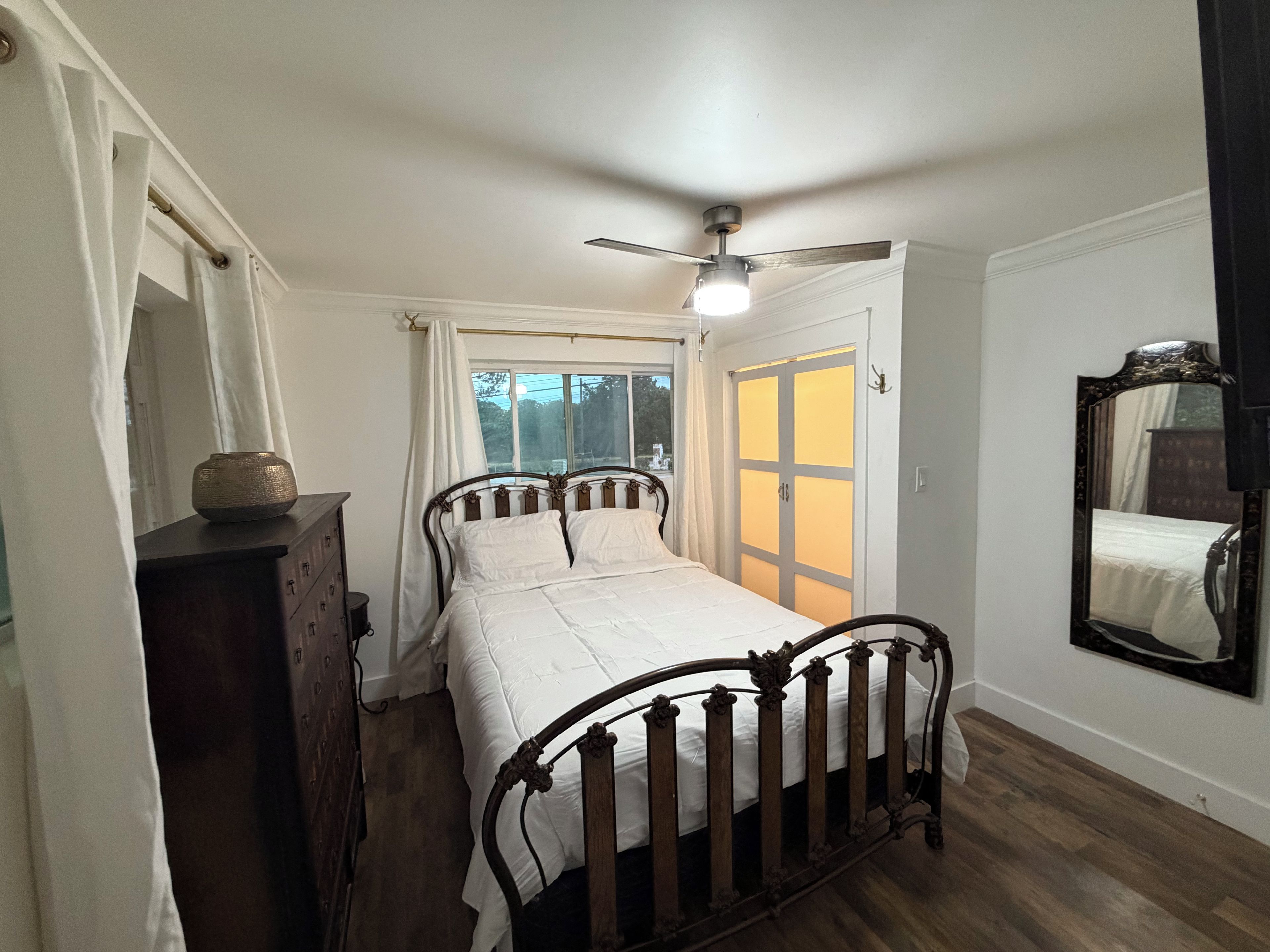 A bedroom with a bed, a dresser, a ceiling fan, a window with curtains, and a mirror on the wall.