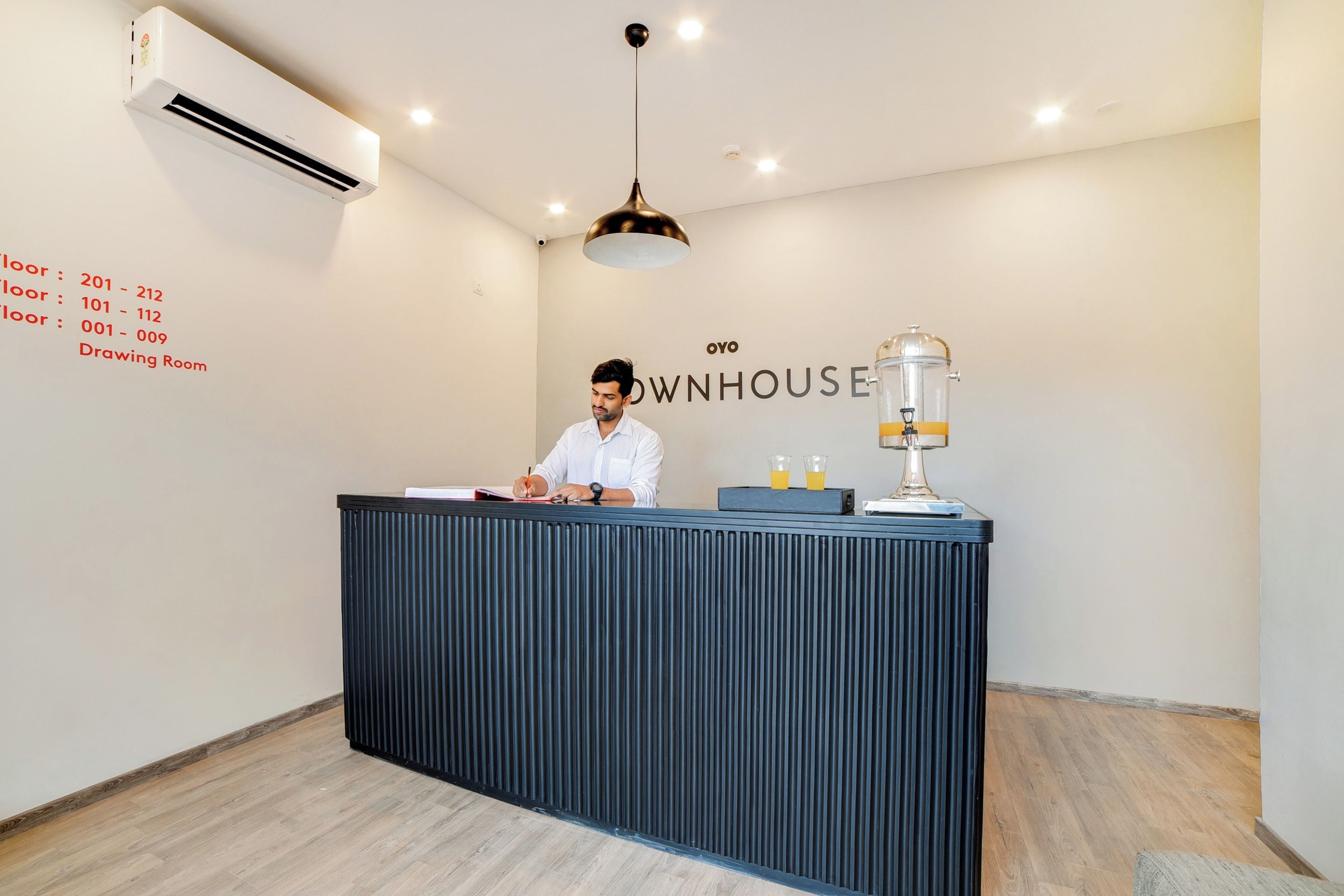 Un mostrador de recepción con una persona sentada detrás, un aire acondicionado de pared y una pared con números de habitaciones y un cartel que dice 'OYO DWNHOUSE'.