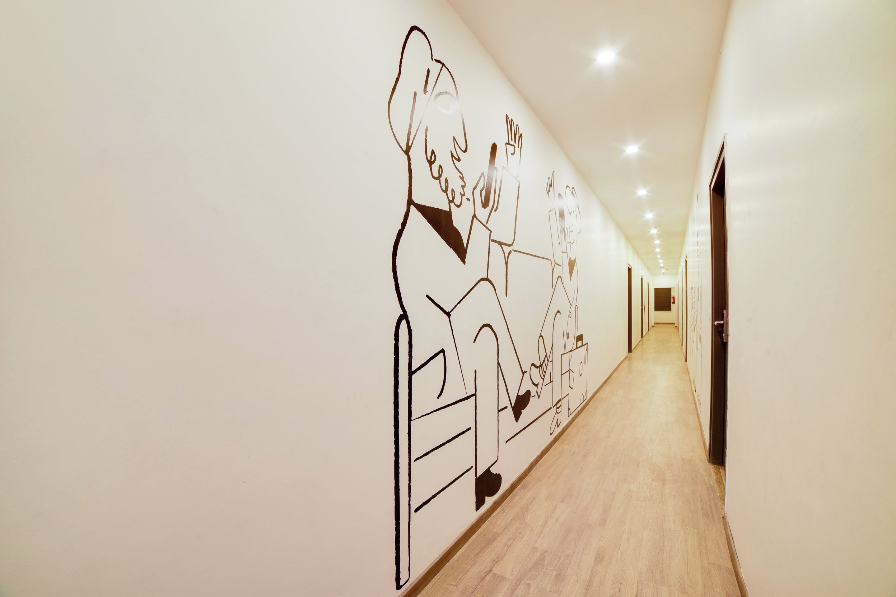 Un pasillo con un mural que muestra a una persona sentada en una mesa con una computadora portátil y otra persona de pie a su lado. El pasillo tiene piso de madera y una serie de puertas a la derecha.