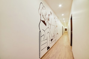 Un pasillo con un mural que muestra a una persona sentada en una mesa con una computadora portátil y otra persona de pie a su lado. El pasillo tiene piso de madera y una serie de puertas a la derecha.