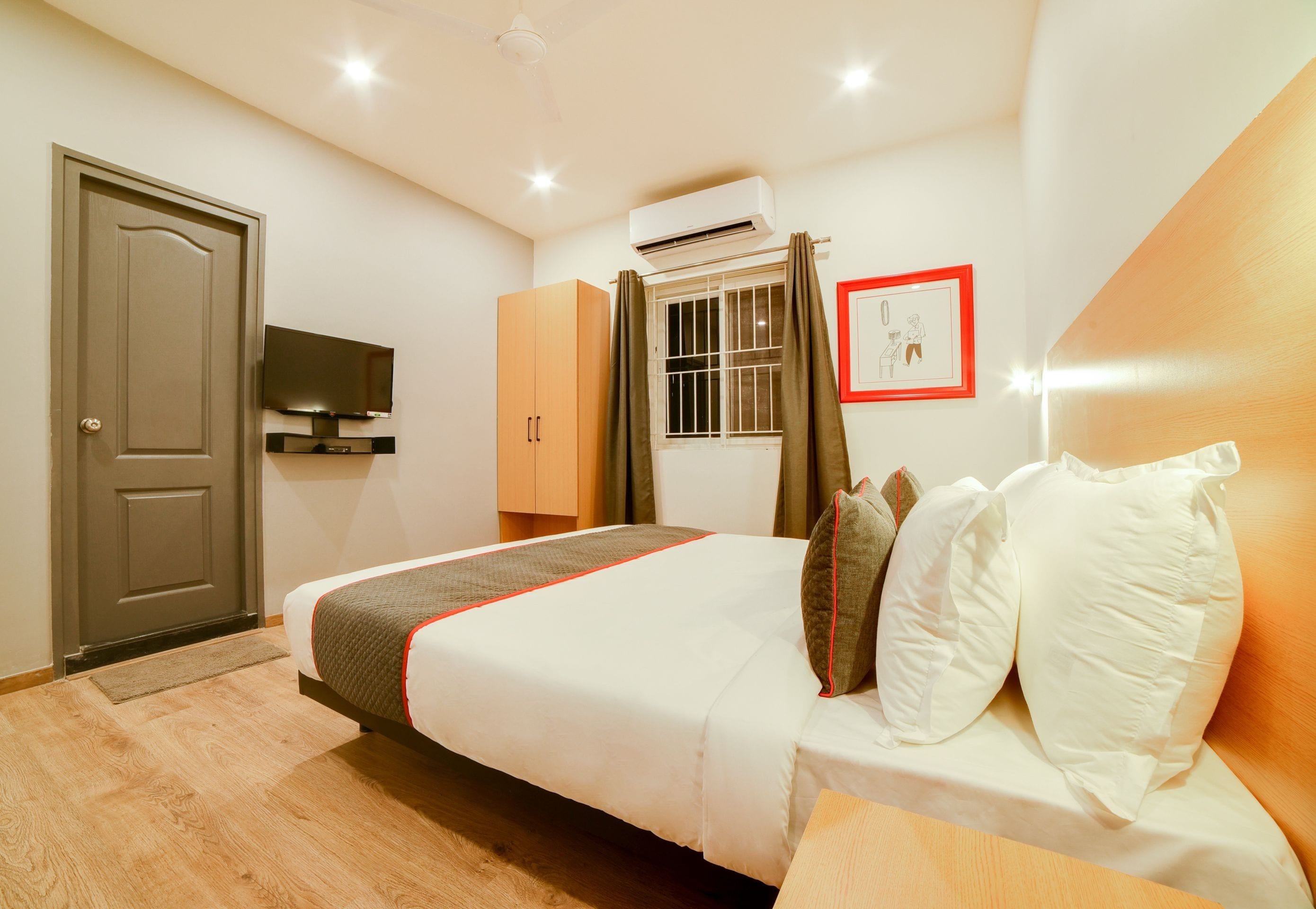 A hotel room with a bed, a television, an air conditioning unit, a window with curtains, and a framed picture on the wall.