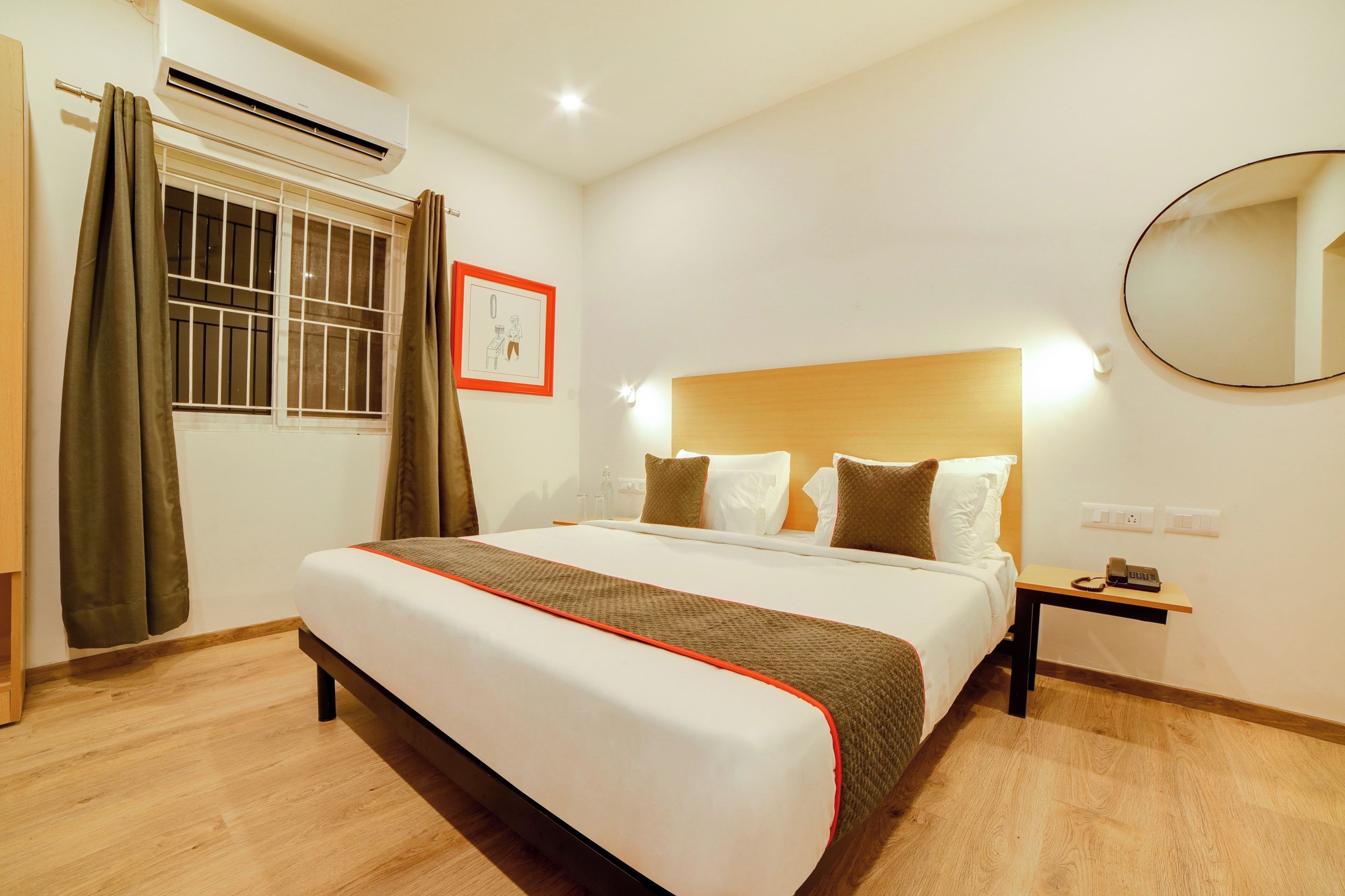 Habitación de hotel con cama, mesita de noche, espejo, ventana con cortinas y aire acondicionado.