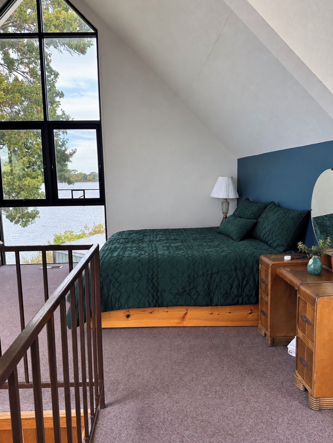 Une chambre avec une grande fenêtre, un lit avec une literie verte, une table de chevet et une commode.