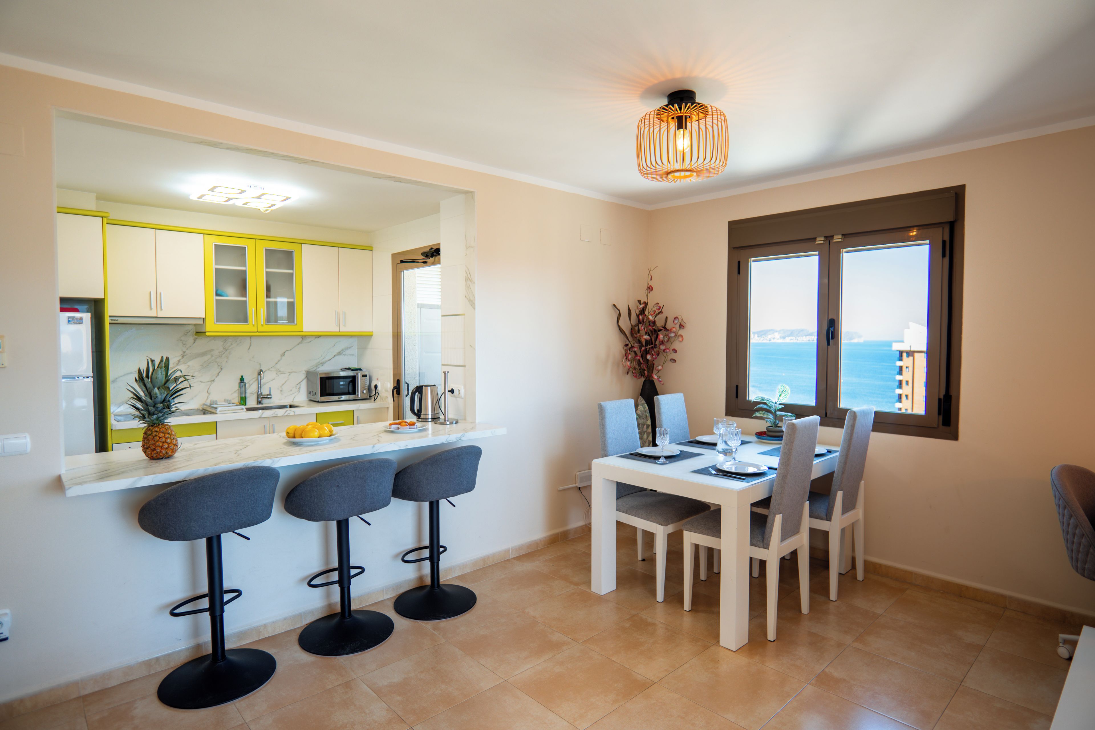 A modern kitchen with a dining area, featuring a large island, bar stools, and a view of the sea through a window.
