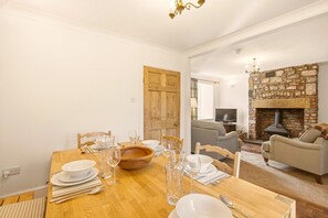 A dining area with a wooden table set for four, a stone fireplace, and a living room with a sofa and television. - Hillside Cottage - North East Escapes (East Ord)