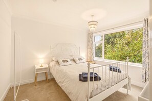 A bedroom with a white bed, a nightstand with a lamp, a window with curtains, and a view of greenery outside. - Hillside Cottage - North East Escapes (East Ord)