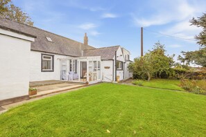 A house with a white exterior, a covered porch, and a well-maintained lawn. - Hillside Cottage - North East Escapes (East Ord)