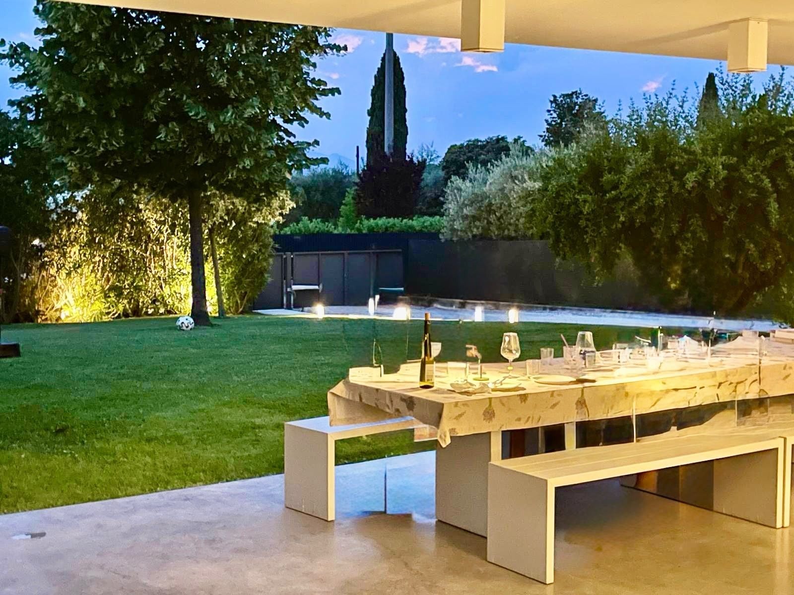 Un espace de restauration en plein air avec une longue table dressée pour un repas, entouré de verdure et offrant une vue lointaine sur le clocher d’une église.