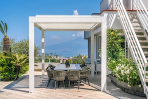 En udendørs terrasse med et bord og stole, en hvid pergola og udsigt til havet og et bjerg.
