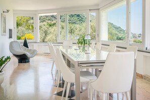 A modern dining area with a glass table and white chairs, large windows with a view of greenery, and a potted plant. - Villa Arpino - Four Bedroom Villa, Sleeps 8 (Vico Equense)