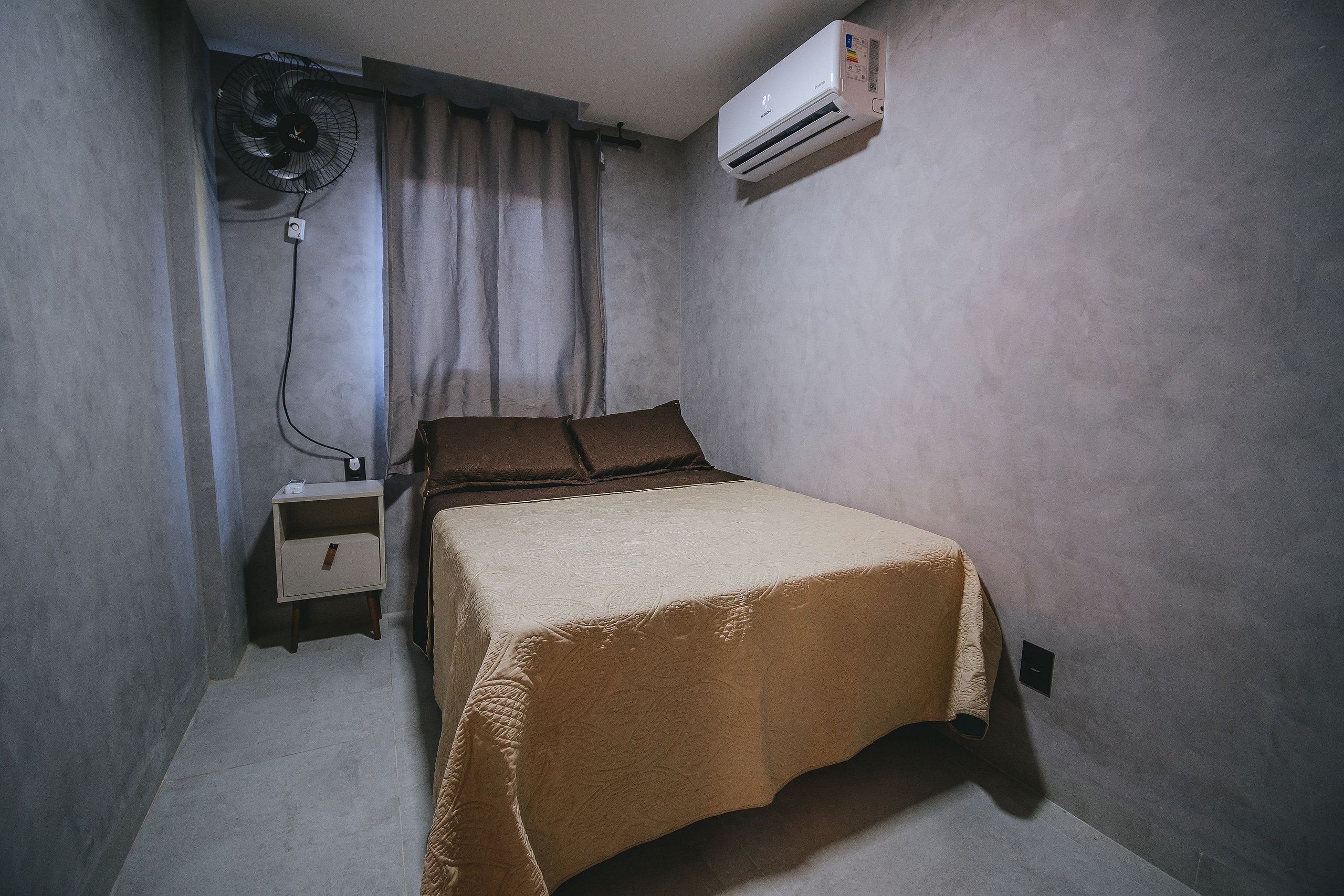 A small, single-bed room with a bed, a bedside table, a wall-mounted air conditioner, and a standing fan.