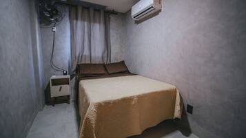 A small, single-bed room with a bed, a bedside table, a wall-mounted air conditioner, and a standing fan.