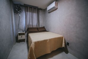 Una habitación pequeña con cama individual, mesita de noche, aire acondicionado de pared y ventilador de pie.