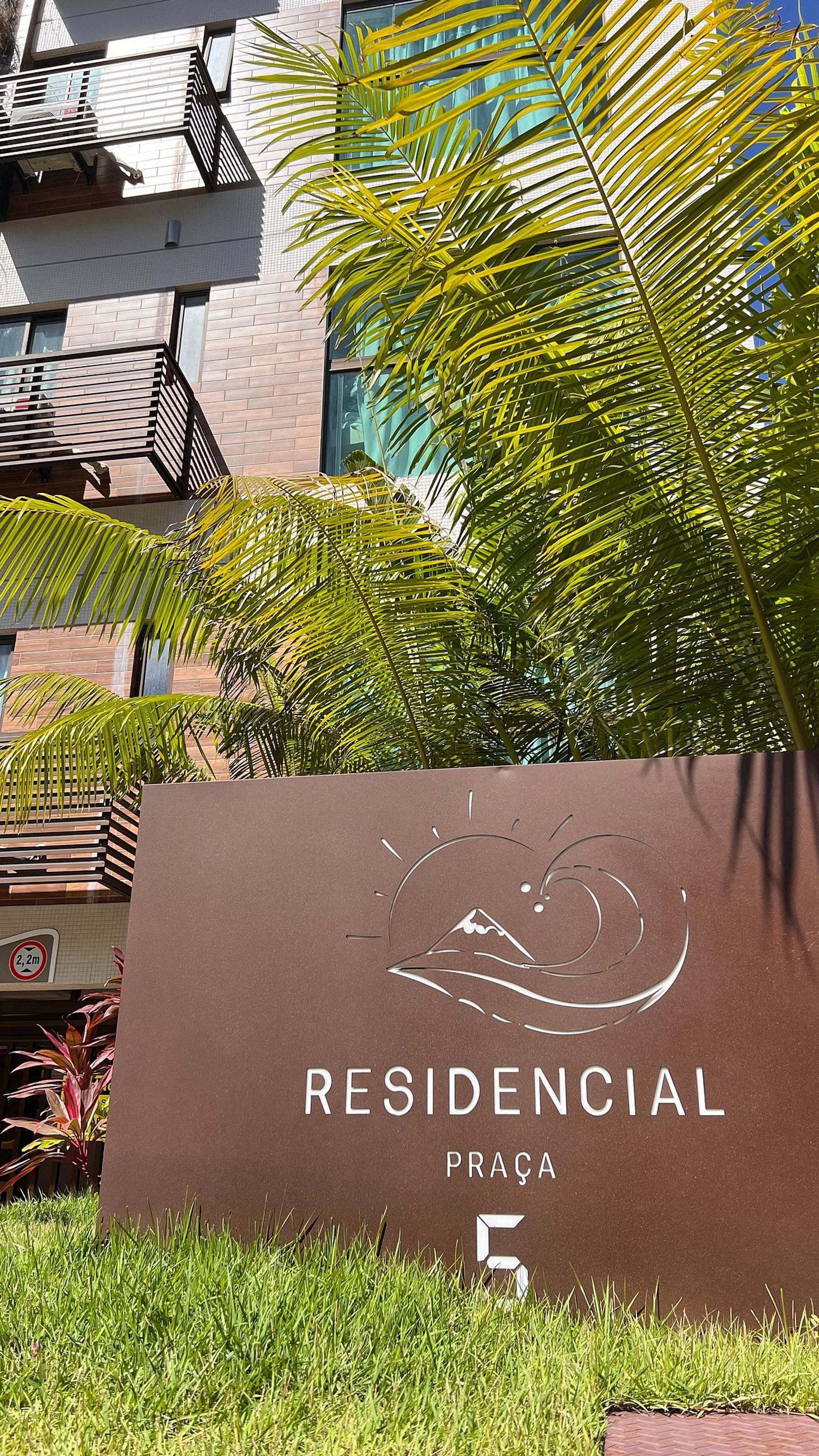 Letrero de 'RESIDENCIAL PRAÇA 5' con diseño estilizado de montaña y ola, rodeado de vegetación y un edificio moderno.