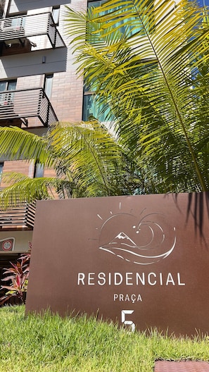 Letrero de 'RESIDENCIAL PRAÇA 5' con diseño estilizado de montaña y ola, rodeado de vegetación y un edificio moderno.