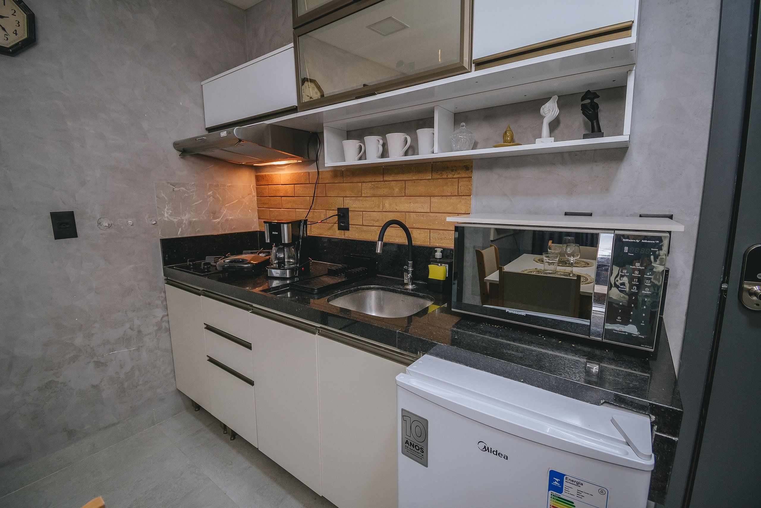 Cocina compacta con armarios blancos, encimera negra, cafetera, microondas y refrigerador.