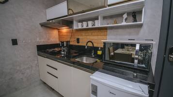 Cocina compacta con armarios blancos, encimera negra, cafetera, microondas y refrigerador.