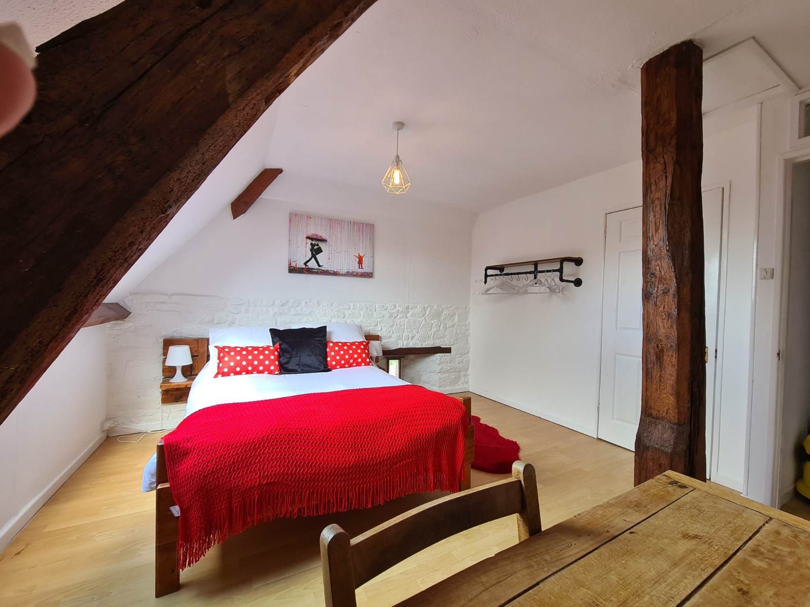 A bedroom with a wooden ceiling, a bed with red and white bedding, a hanging light fixture, and a white wall with a picture.