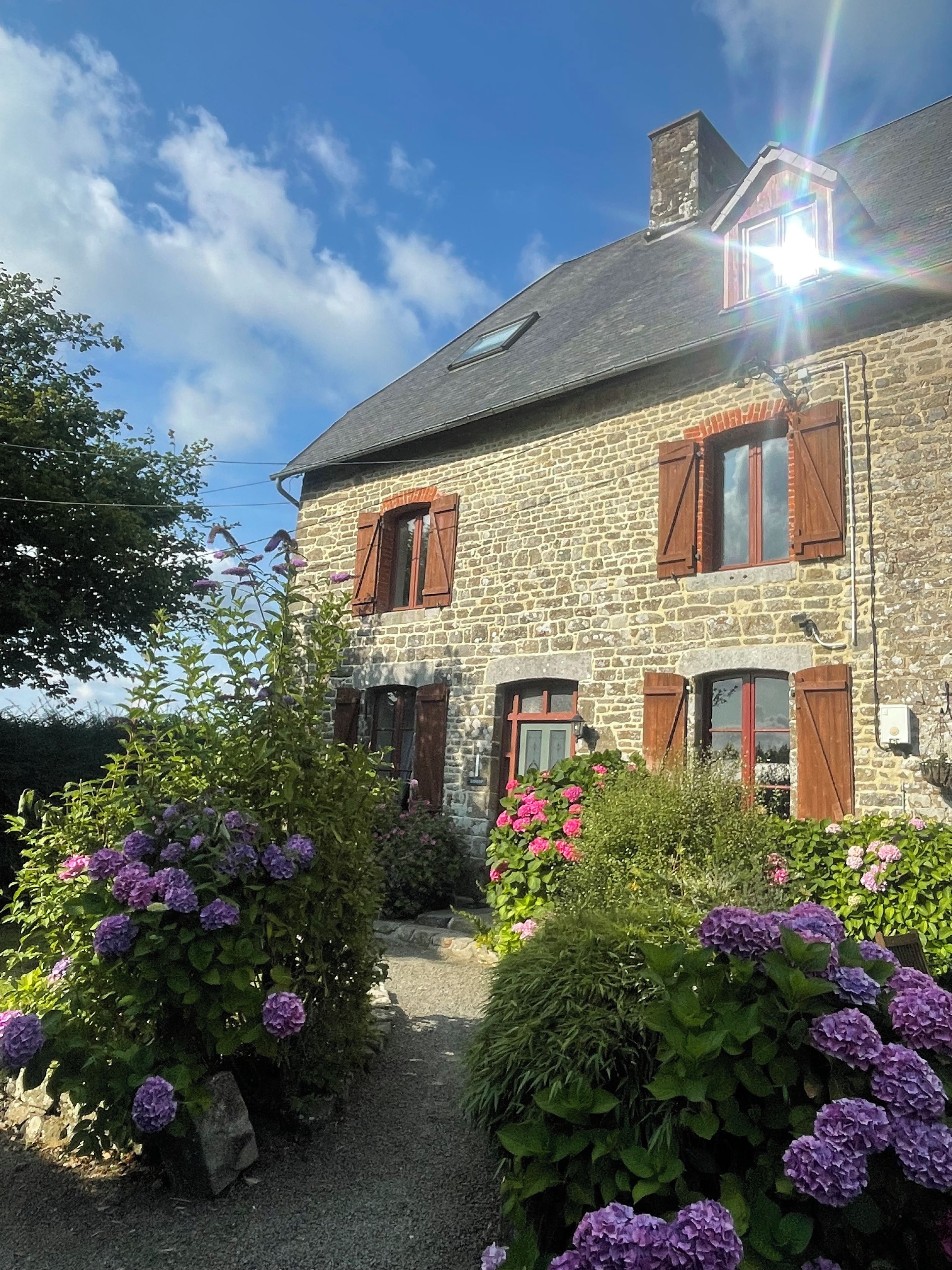 A stone building with multiple windows and shutters, surrounded by lush greenery and flowering plants.
