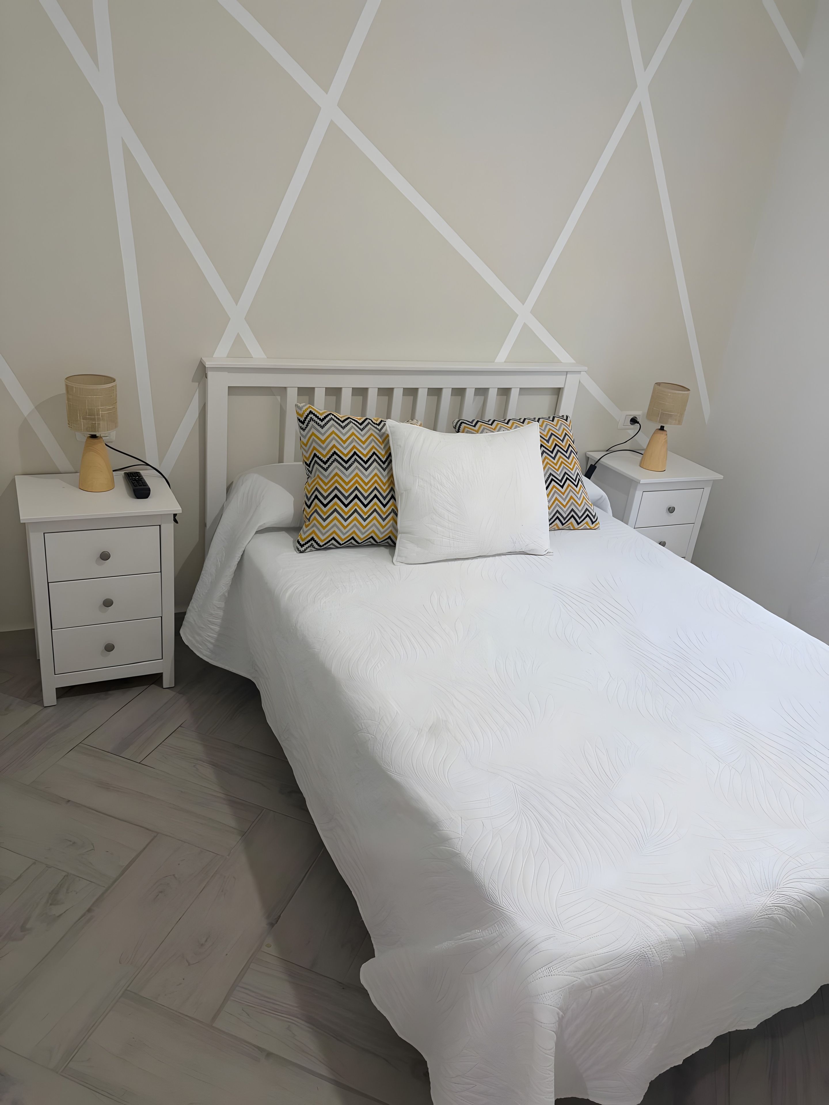 A neatly made bed with a white bedspread and two patterned pillows, flanked by white nightstands with lamps and a remote control.
