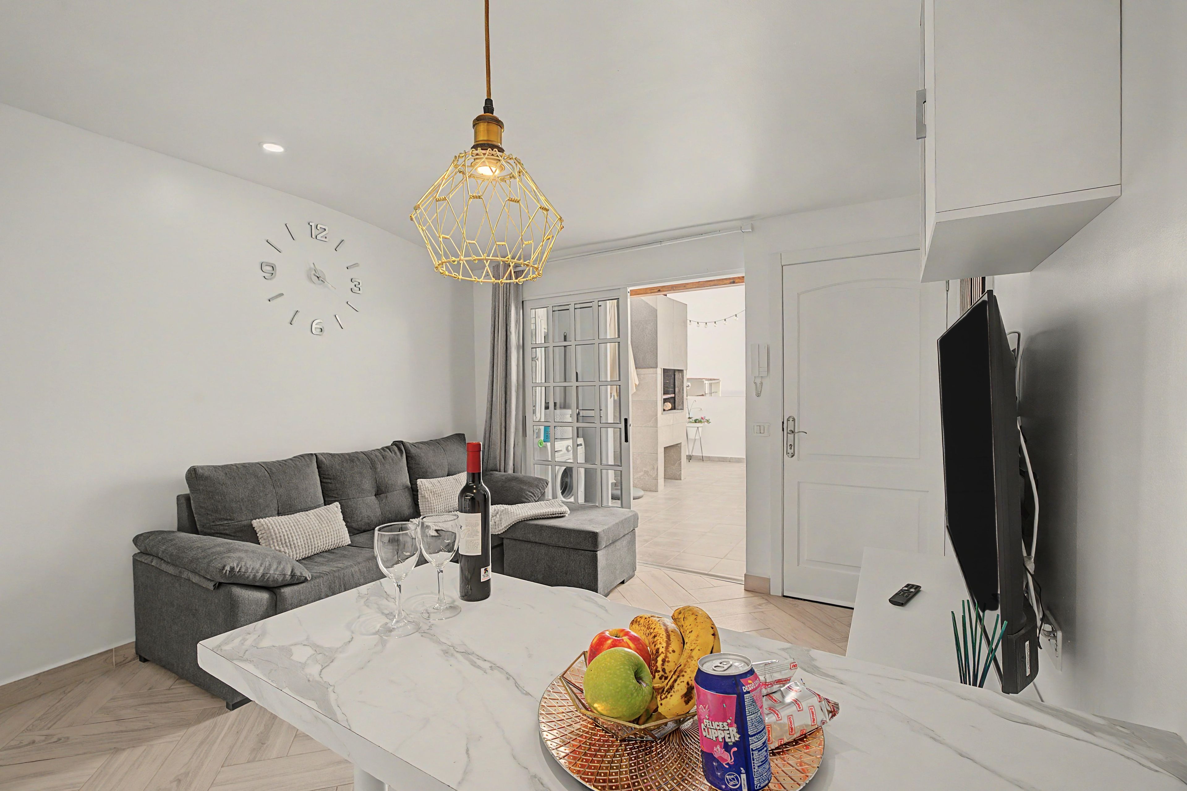 A modern living room with a grey sofa, a marble coffee table, and a dining area with a fruit bowl and beverages.