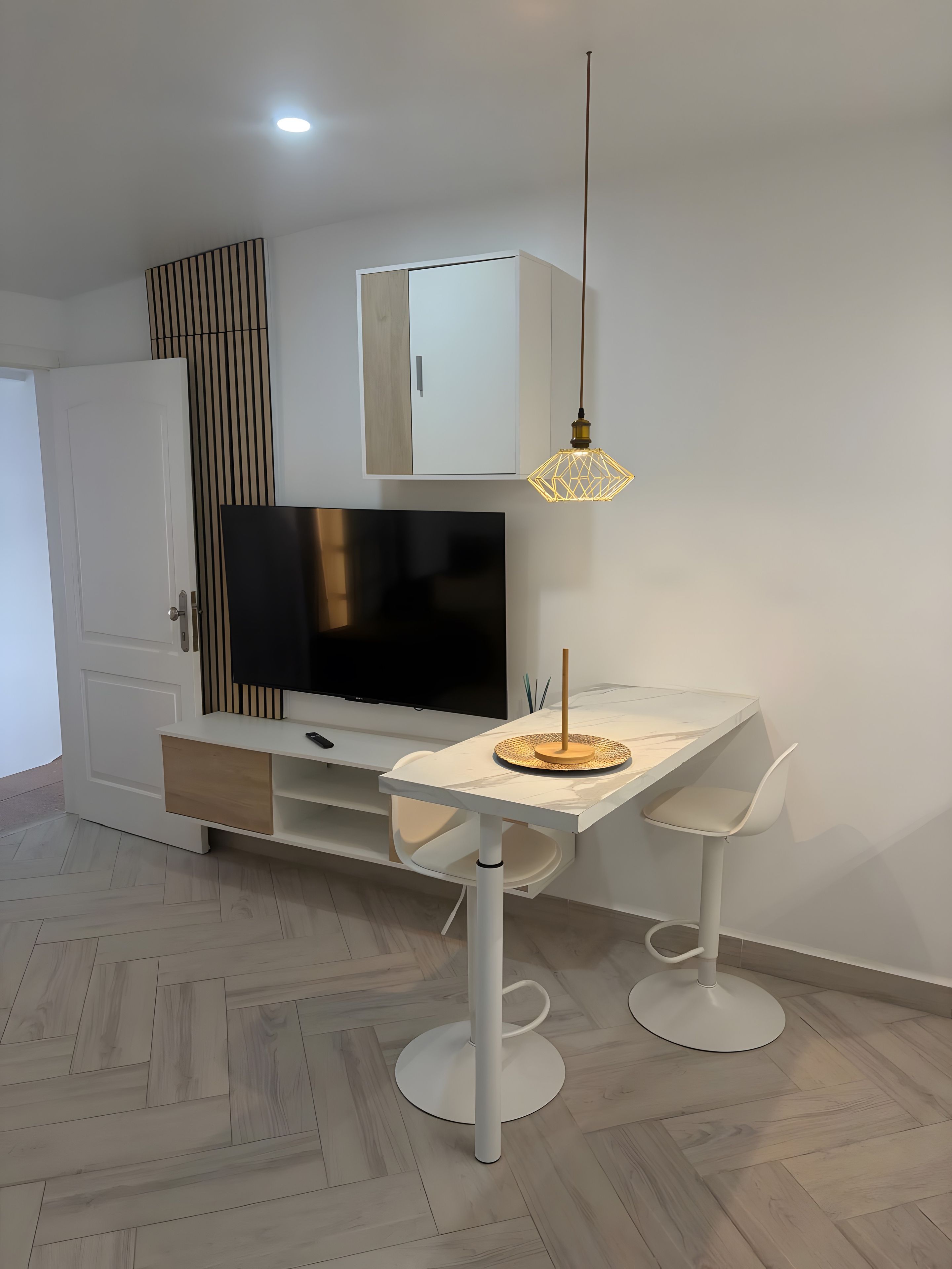 Una sala de estar moderna con un televisor de pantalla plana, un armario blanco, una mesa de comedor con dos sillas y una lámpara colgante.