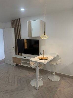 Una sala de estar moderna con un televisor de pantalla plana, un armario blanco, una mesa de comedor con dos sillas y una lámpara colgante.
