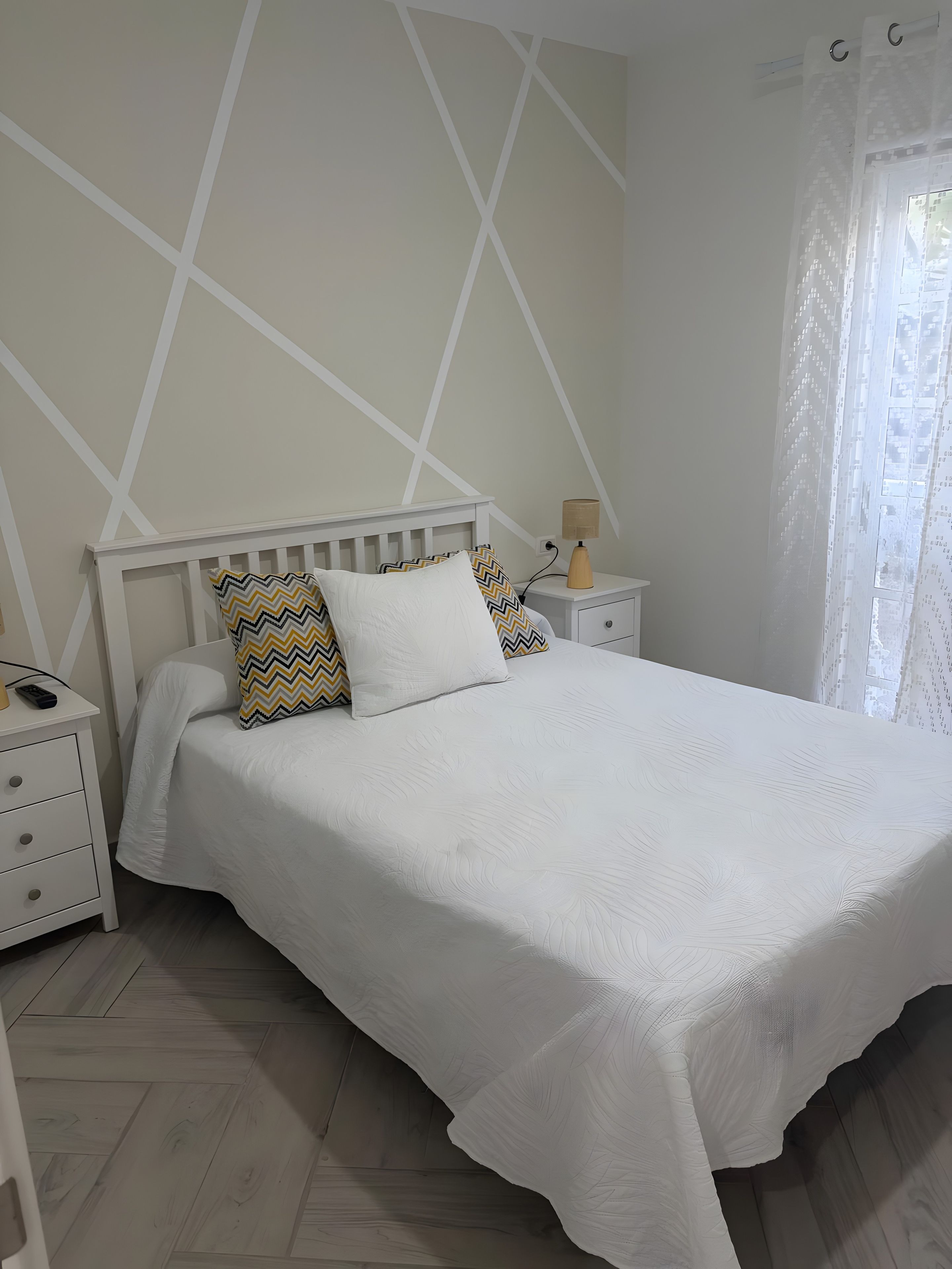 Un dormitorio con una cama blanca, una mesita de noche, una lámpara y una ventana con cortina.