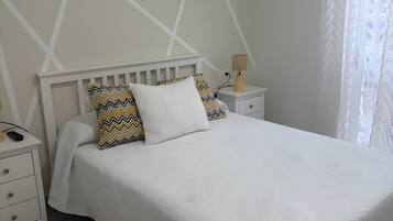 Un dormitorio con una cama blanca, una mesita de noche, una lámpara y una ventana con cortina.
