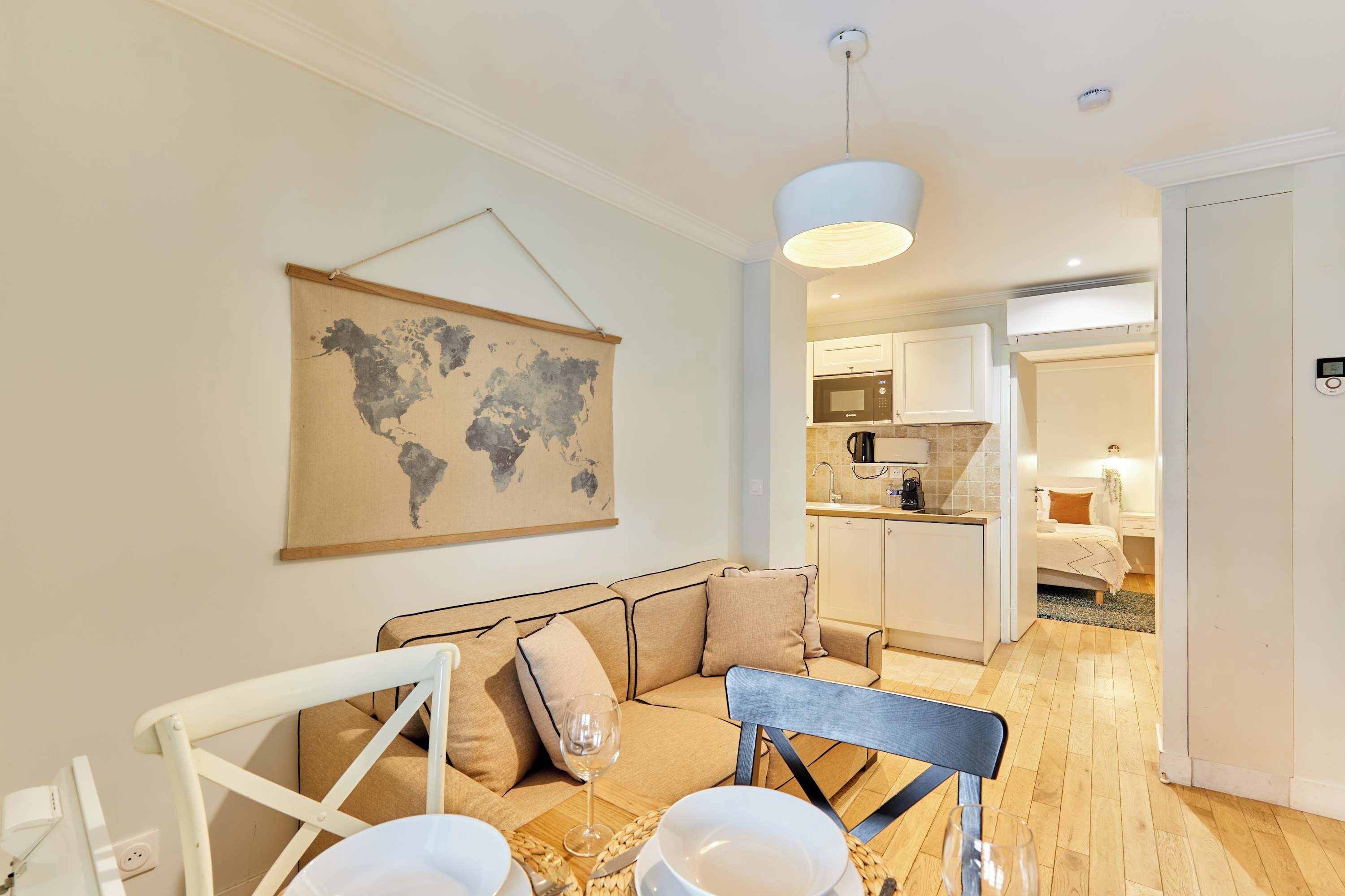 A modern living room with a sofa, a dining table with chairs, a hanging world map, a kitchen in the background, and a hallway leading to a bedroom.