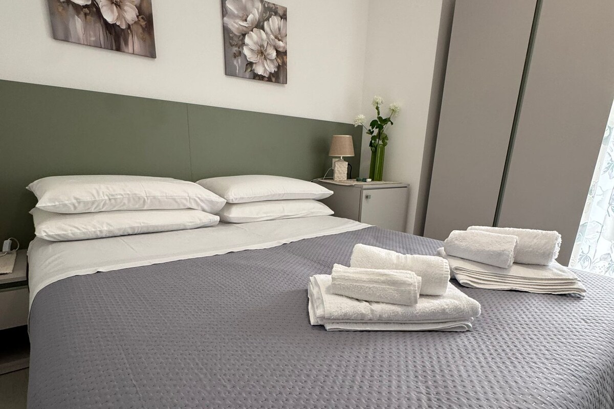 A neatly made bed with white pillows and a grey quilt, a bedside table with a lamp and a vase of flowers, and two floral paintings on the wall.