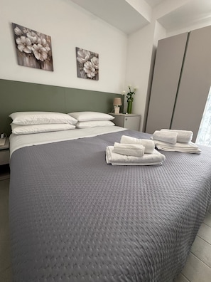 A neatly made bed with white pillows and a grey quilt, a bedside table with a lamp and a vase of flowers, and two floral paintings on the wall. - A un Passo dalla Piazza (San Giovanni Gemini)