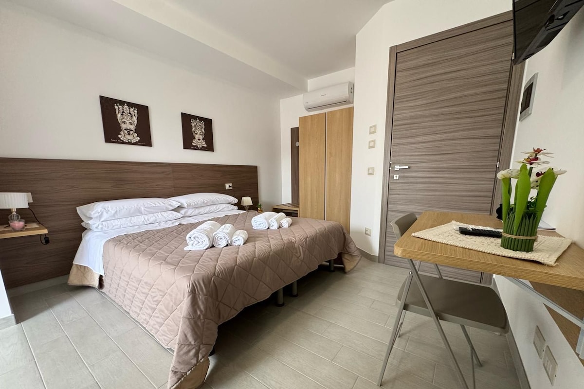A hotel room with a bed, bedside table, lamp, air conditioning unit, wardrobe, and a small desk with a vase of flowers.