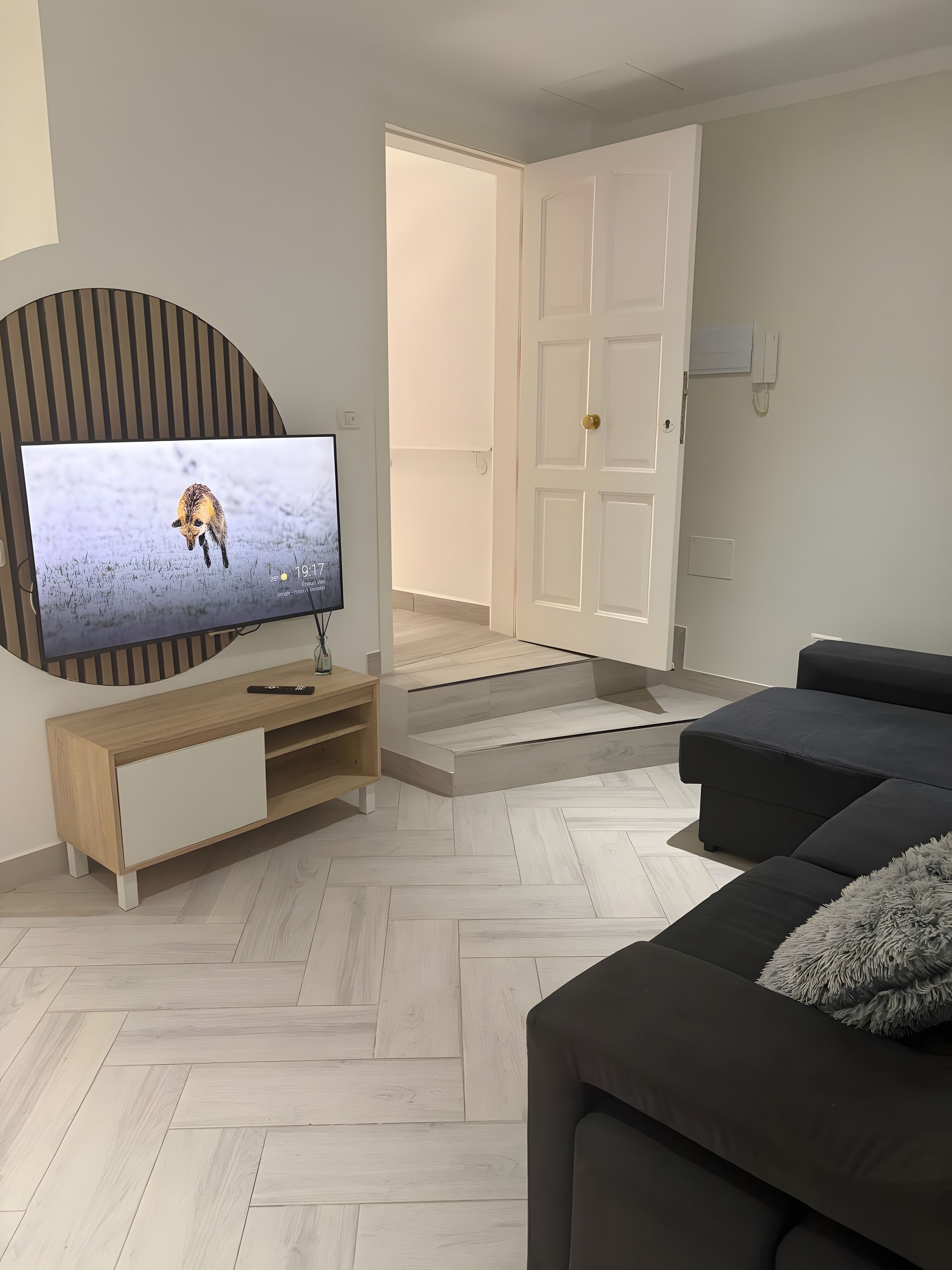 A modern living room with a flat-screen TV on a wooden console, a dark sofa, and a herringbone-patterned floor.